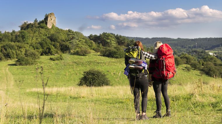 Zwei Wanderer mit Rucksäcken stehen auf einer grünen Wiese. Im Hintergrund ist eine Burgruine und ein blauer Himmel zu sehen.