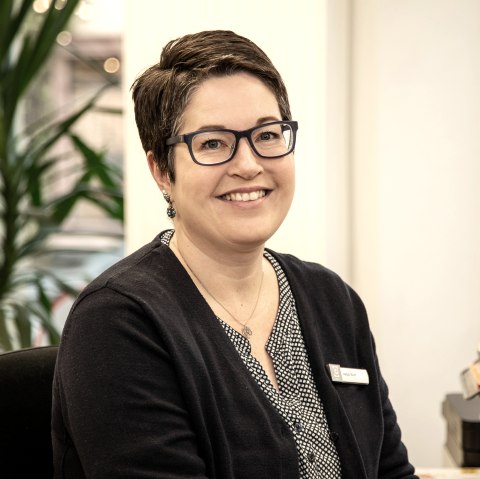 A woman sits at a desk and smiles at the camera. Behind her is a large palm tree.