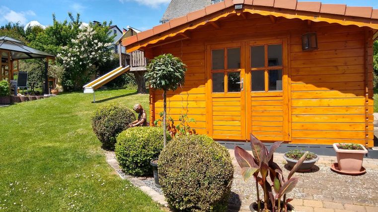 Ein gemütliches, Holzhaus im Garten mit gepflegten Zierpflanzen. Im Hintergrund sind grüne Wiesen und ein blauer Himmel zu sehen.