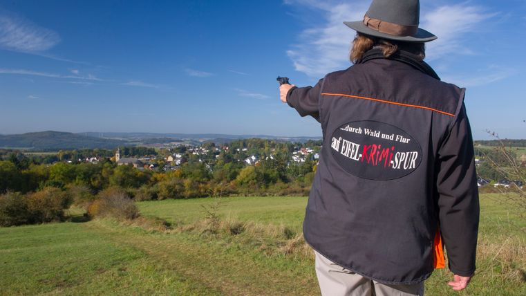 Person mit Hut und Jacke mit Aufschrift 'auf EIFEL KRIMI SPUR' zeigt auf eine Landschaft mit Dorf im Hintergrund.