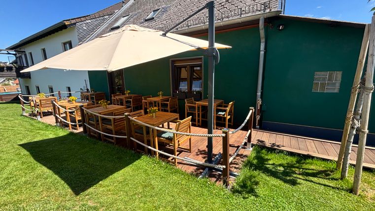 Eine gemütliche Außenterrasse mit Holzstühlen und Tischen. Ein Sonnenschirm bietet Schatten und die Umgebung ist grün und einladend.