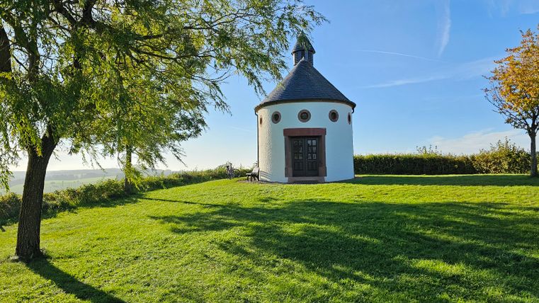 Runde Kapelle auf grüner Wiese mit Bäumen und blauem Himmel.