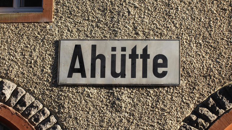 Ein Bahnhofsschild mit der Aufschrift 'Ahütte' hängt an einer grob verputzten Wand neben einem gemauerten Bogen.