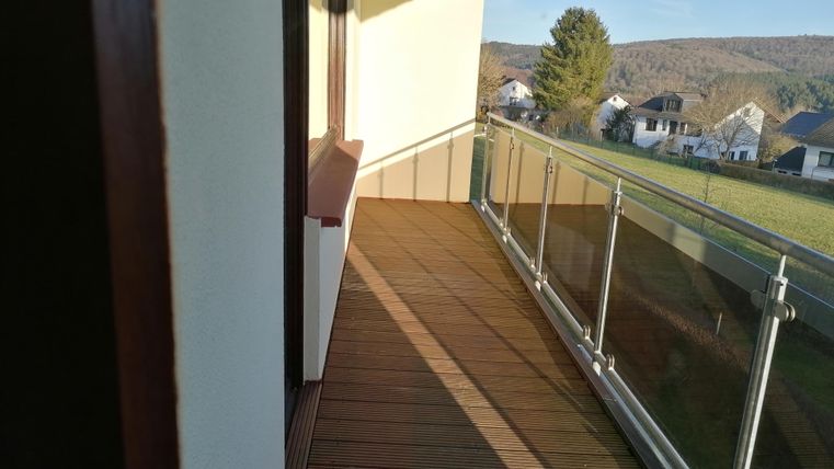 A sunny balcony with a wooden floor and a glass balustrade. In the background, green meadows and some residential houses can be seen.