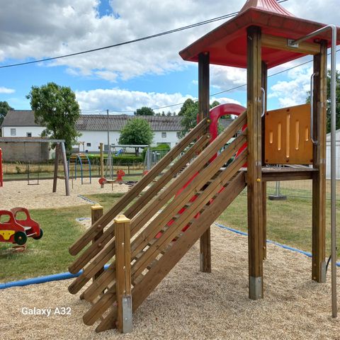 Spielplatz mit Klettergerüst und Rutsche, umgeben von Schaukeln und Wippen. Im Hintergrund sind Bäume und Gebäude zu sehen. Der Himmel ist teils bewölkt.