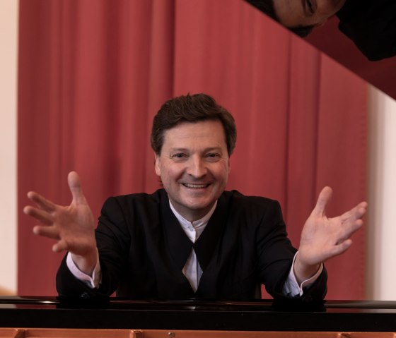Ein Mann sitz an einem Flügel und schaut mit ausgebreiteten Armen lachend in die Kamera