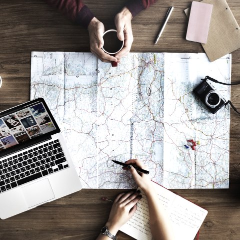 Large wooden table with two people sitting at it. On it a map, cameras, notes, pens and a laptop.