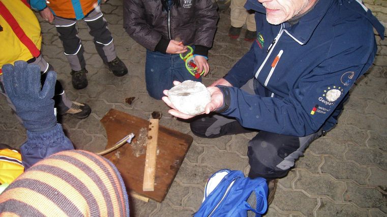 Eine Kindergruppe steht um einen Mann herum, welcher Ihnen einen Stein zeigt und etwas erklärt.