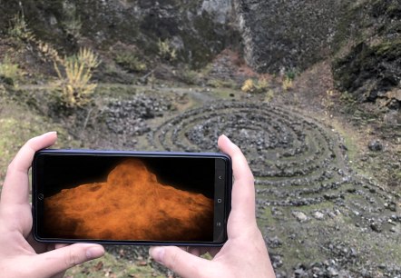 Two hands hold a smartphone across the abyss of a volcano. The volcanic explosion is visualised on the screen.