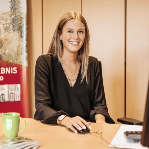 A woman sits at a desk and smiles at the camera. Behind her are some cupboards and a banner with the words ‘Freizeirerlebnis Gerolsteiner Land’ (Gerolsteiner Land leisure experience) and a picture of a castle.