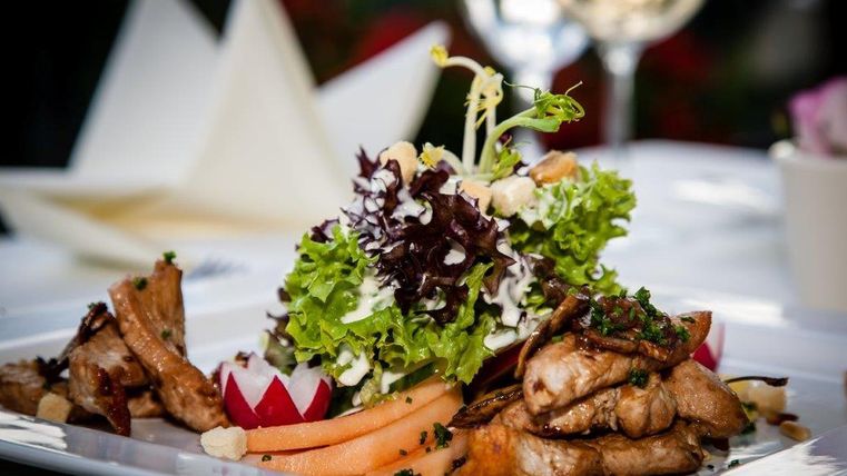 Ein kunstvoll angerichtetes Gericht mit Fleisch, Salat, Radieschen und Melone auf einem weißen Teller, im Hintergrund ein Glas Weißwein.
