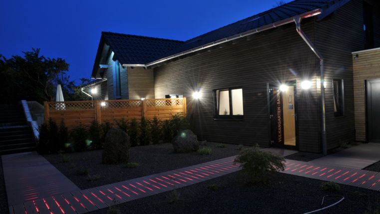 Une maison moderne avec une façade en bois et une entrée illuminée la nuit. Le chemin menant à la maison est élégamment marqué par des lumières rouges.
