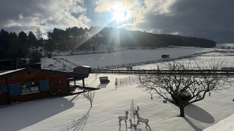 Ein winterlicher Landschaft mit viel Schnee und einem strahlenden Sonnenlicht. Im Vordergrund ist ein Holzhaus und einige dekorative Figuren zu sehen.