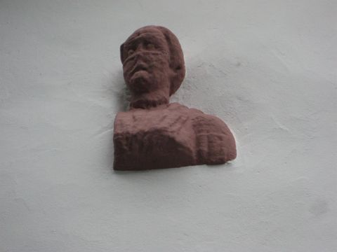 Aus einer weißen Wand schaut ein Torso eines Mannes hervor.Der aus rötlichem Stein bestehene Statue besteht aus Kopf und Brust und stammt aus der Römerzeit.