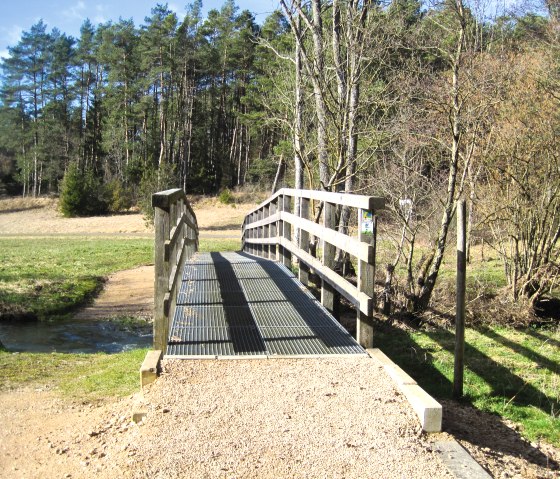 Kleine Holzbr&uuml;cke &uuml;berquert den Nohner Bach in einer l&auml;ndlichen Landschaft mit B&auml;umen und blauem Himmel., &copy; Touristik GmbH Gerolsteiner Land, Ute Klinkhammer