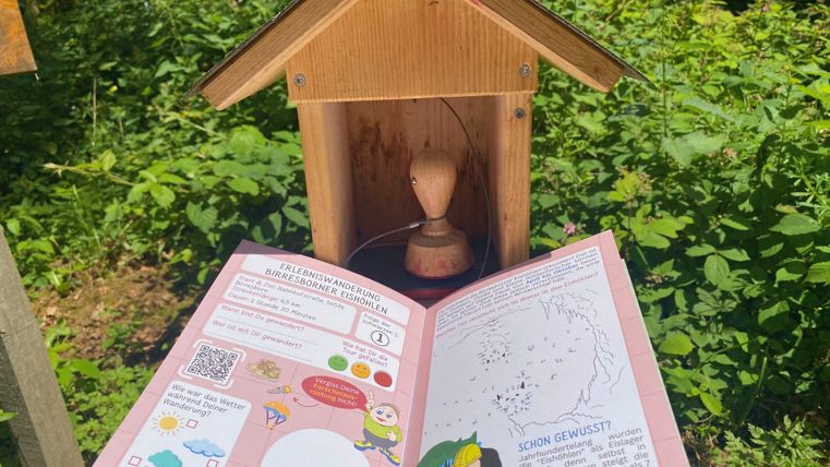 Geöffnetes Prospekt "Wander(S)pass für Kids" mit Rätselseite wird hochgehalten vor der dazugehörigen Stempelstation.