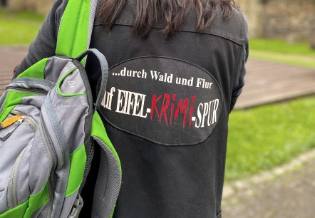 A person wearing a hat stands in front of a large town wall with her back to the viewer. She is wearing a jacket with the inscription "...through woods and meadows on the Eifel crime trail" on the back and is carrying a rucksack over one shoulder.