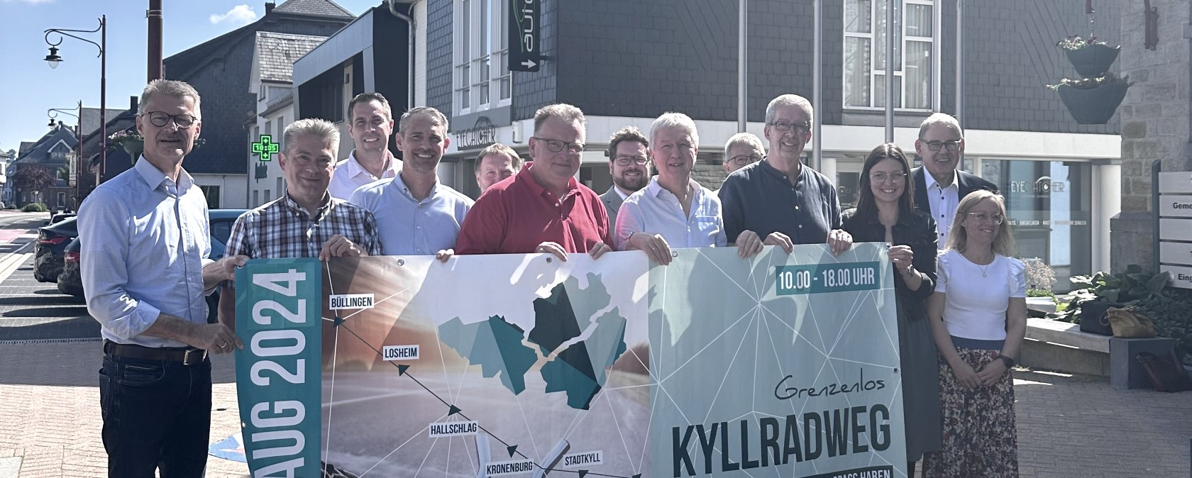 13 Personen stehen dicht beieinander und halten ein Veranstaltungsbanner f&uuml;r den Radaktionstga Grenzenlos kyllradweg am 11. August 2024 hoch.