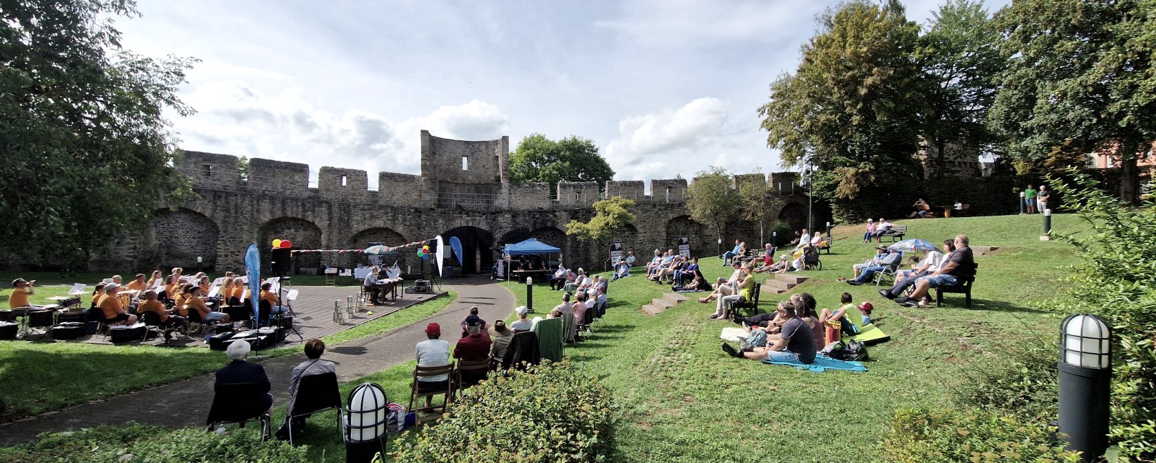 Zahlreiche Personen sitzen auf Stühlen und Decken auf einer Wiese im Park und schauen zur kleinen Bühne mit Musikverein und Lesern vor der Hillesheimer Stadtmauer