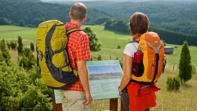 Deux randonneurs avec des sacs à dos se tiennent devant un point de vue et regardent le paysage. À l'arrière-plan, on aperçoit des collines douces et des forêts.