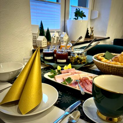 Ein schön gedeckter Tisch mit goldenen Servietten, Schalen und einer Auswahl an frischen Lebensmitteln. Im Hintergrund sind dekorative kleine Häuschen und Pflanzen zu sehen.